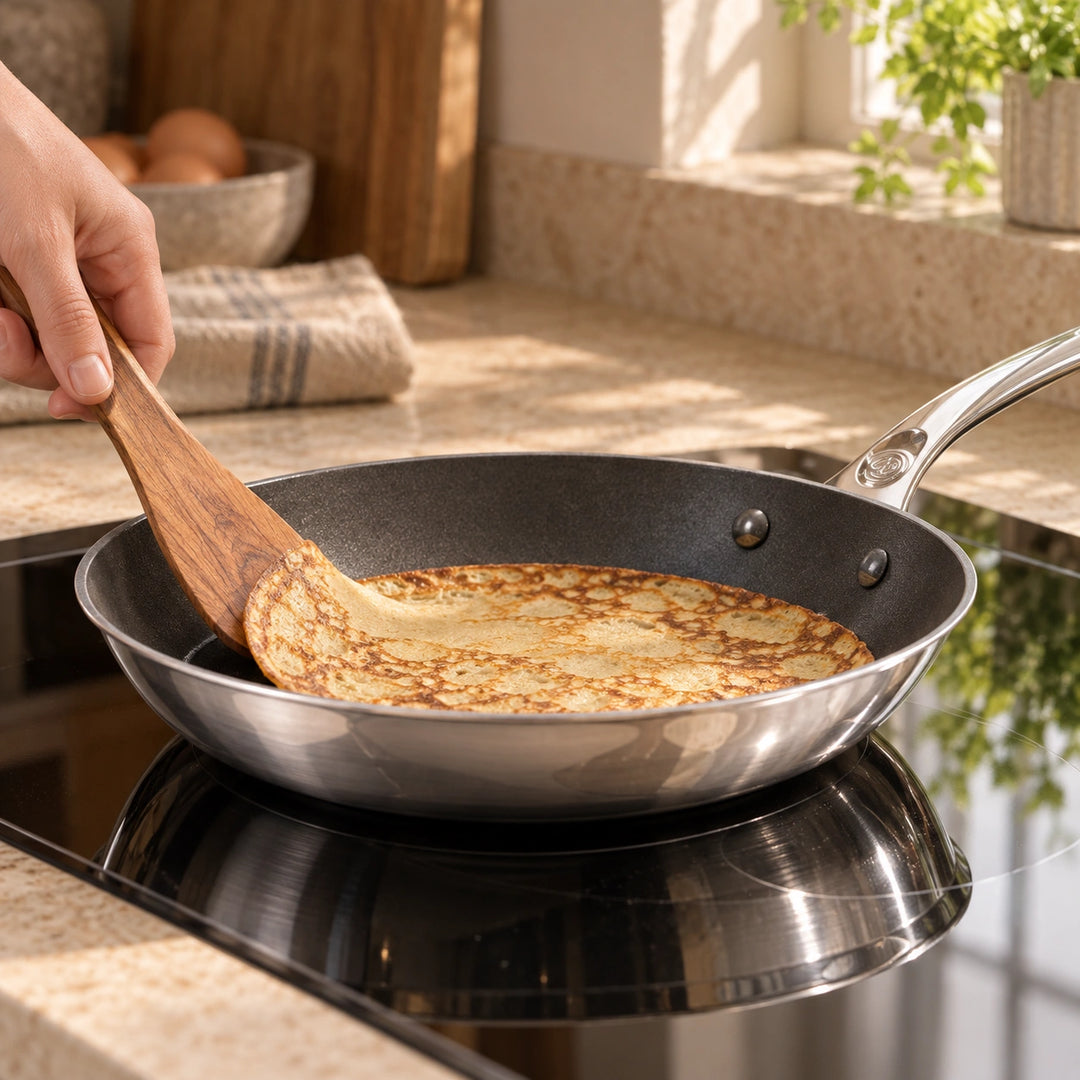 Anwendungsbeispiel: Eine Hand hebt einen goldbraunen Crêpe mit einem Holzspatel in der beschichteten Pfanne Affinity an.
