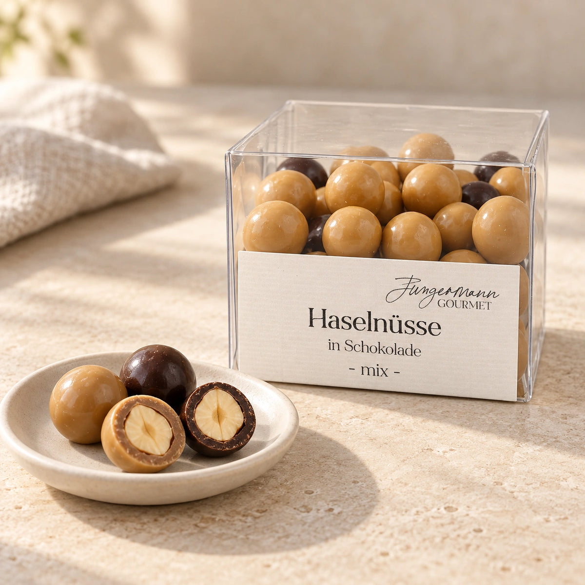 Haselnüsse in Schokolade mix in der Originalbox neben ganzen und halbierten Schokoladenhaselnüssen auf einer kleinen Keramikschale.