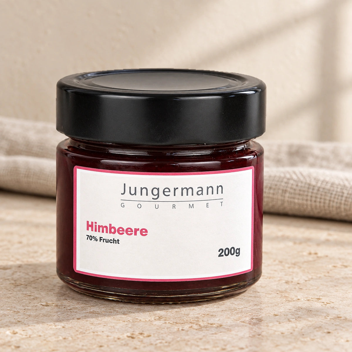 Himbeer-Aufstrich im 200-g-Originalglas mit schwarzem Deckel auf hellem Stein vor einem Leinentuch.