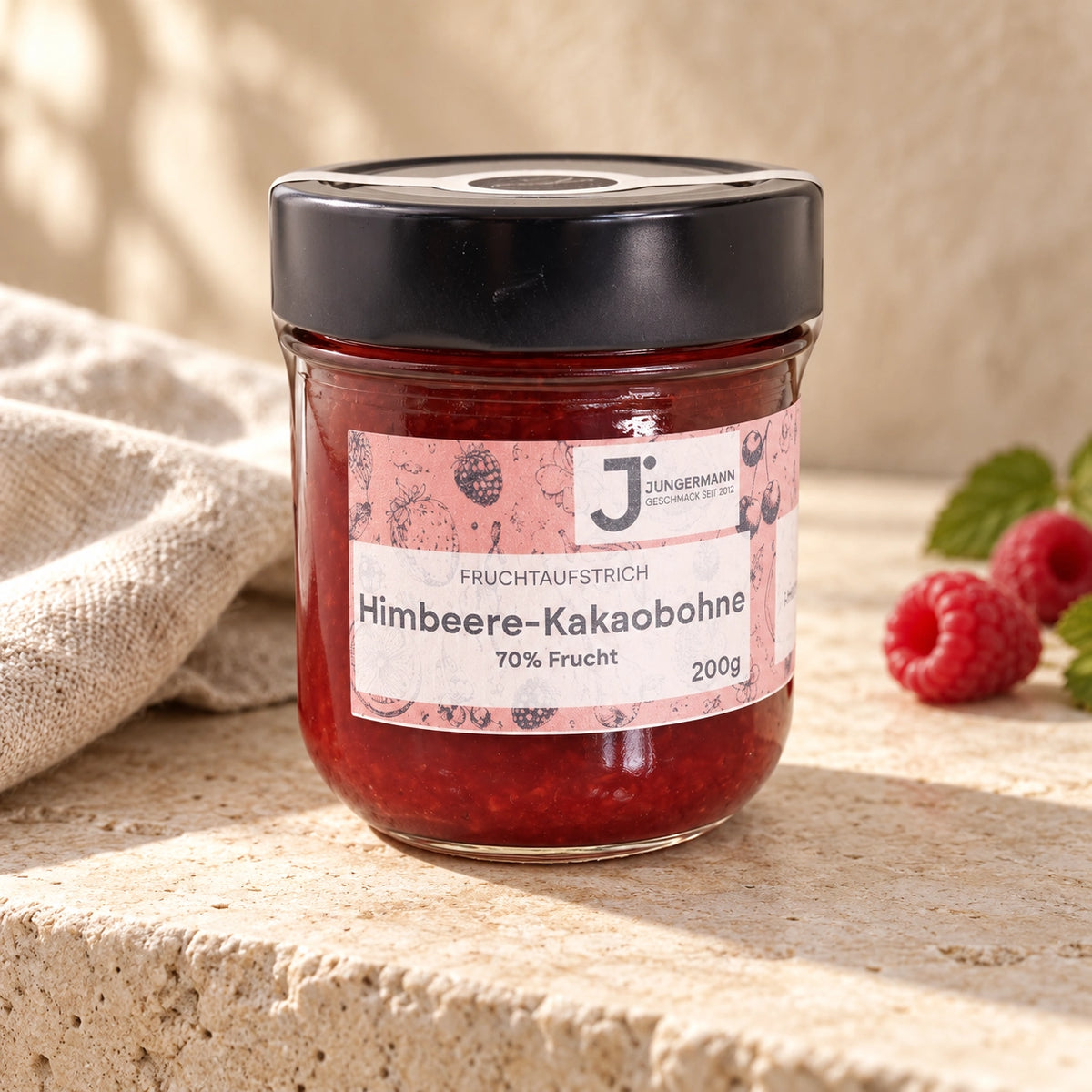 Himbeere-Kakaobohne-Aufstrich im Glas mit rosa Etikett auf hellem Naturstein neben einem Leinentuch.