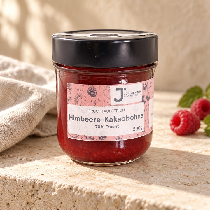 Himbeere-Kakaobohne-Aufstrich im Glas mit rosa Etikett auf hellem Naturstein neben einem Leinentuch.