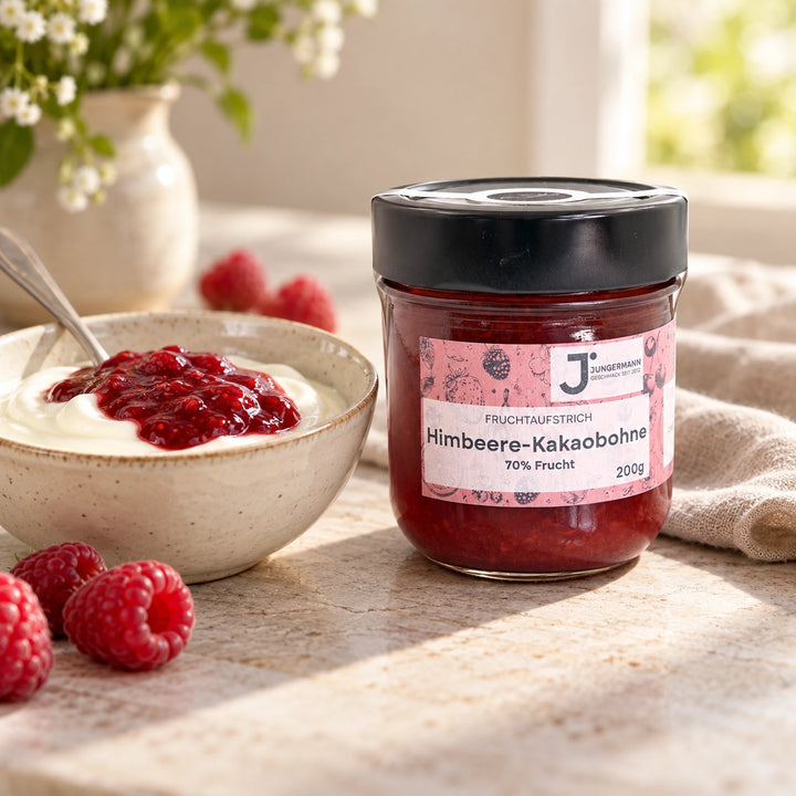 Himbeere-Kakaobohne-Aufstrich im Originalglas neben einer Schale Joghurt mit rotem Fruchtaufstrich als Serviervorschlag.
