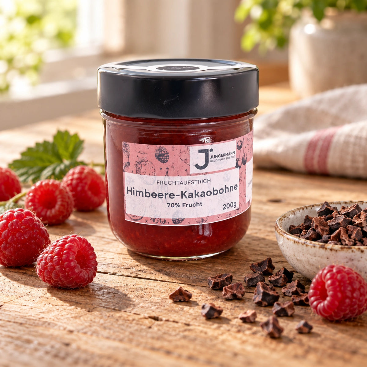 Himbeere-Kakaobohne-Aufstrich im erkennbaren Originalglas neben Himbeeren und einer kleinen Schale Kakaobohnensplitter.