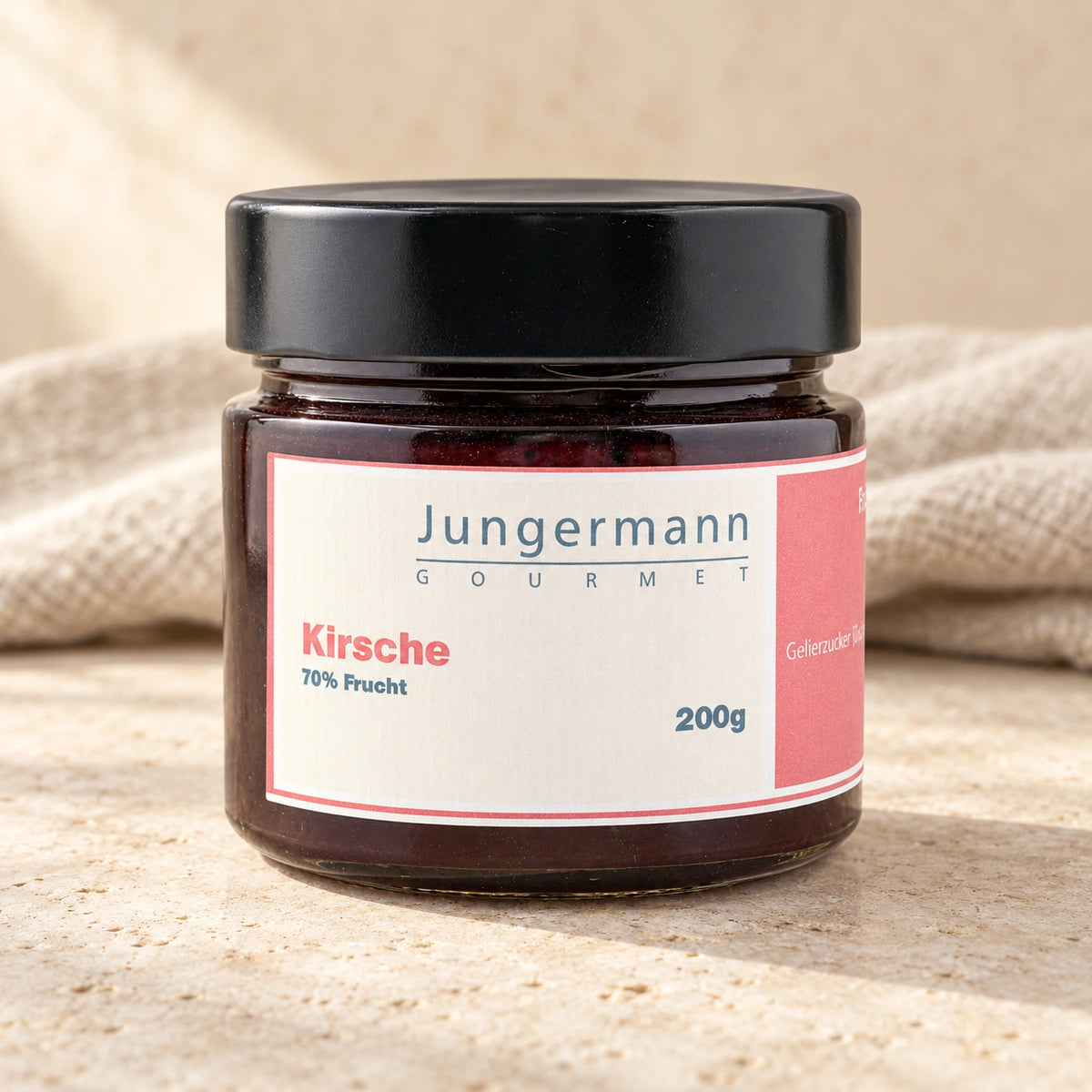 Kirsch-Aufstrich im 200-g-Originalglas mit schwarzem Deckel auf hellem Naturstein vor einem Leinentuch.