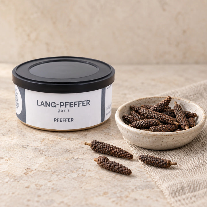 Langpfeffer in der Originaldose neben einer Keramikschale mit ganzen getrockneten Pfefferähren auf hellem Stein.