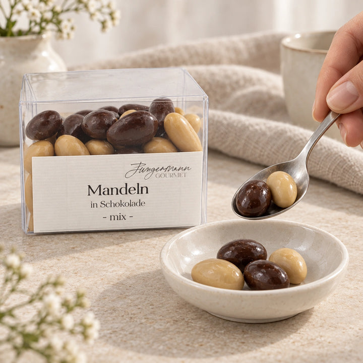 Eine Hand portioniert helle und dunkle Schokoladenmandeln mit einem Löffel in eine Keramikschale; daneben steht die Originalbox Mandeln in Schokolade Mix von Jungermann Gourmet