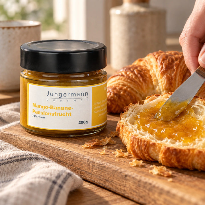 Ein Croissant wird mit Mango-Banane-Passionsfrucht-Aufstrich bestrichen; daneben steht das Originalglas. Serviervorschlag.