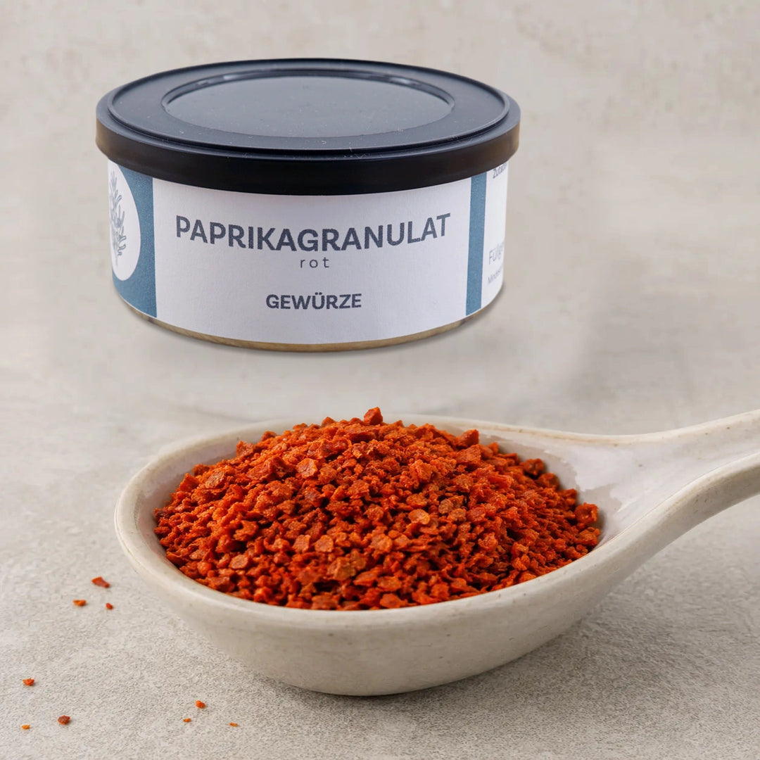 Rotes Paprikagranulat auf einem Keramiklöffel vor der vollständig sichtbaren Originaldose.