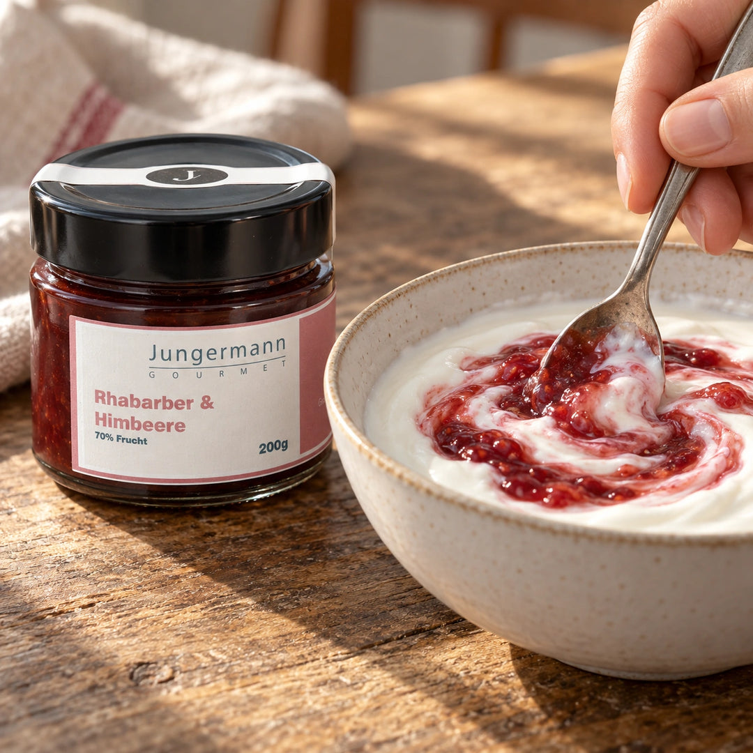 Eine Hand rührt Rhabarber-Himbeer-Aufstrich in Naturjoghurt; das Originalglas steht daneben. Serviervorschlag.