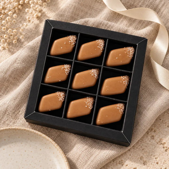 Neun Salz-Karamell-Pralinen in der schwarzen Originalbox mit neun Fächern auf Leinen, daneben ein loses Dekoband.