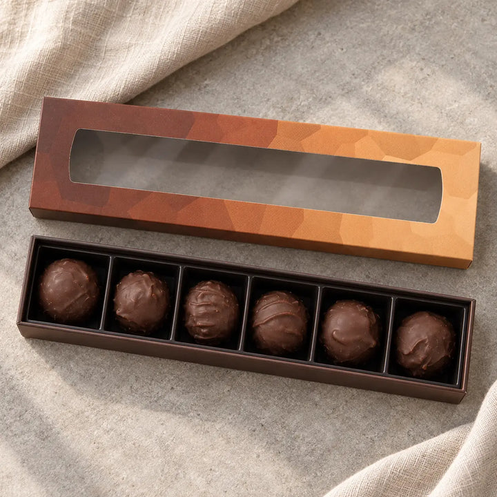 Geöffnete Originalbox mit genau sechs Schokoladen Trüffeln in einer Reihe im schwarzen Fächereinsatz.