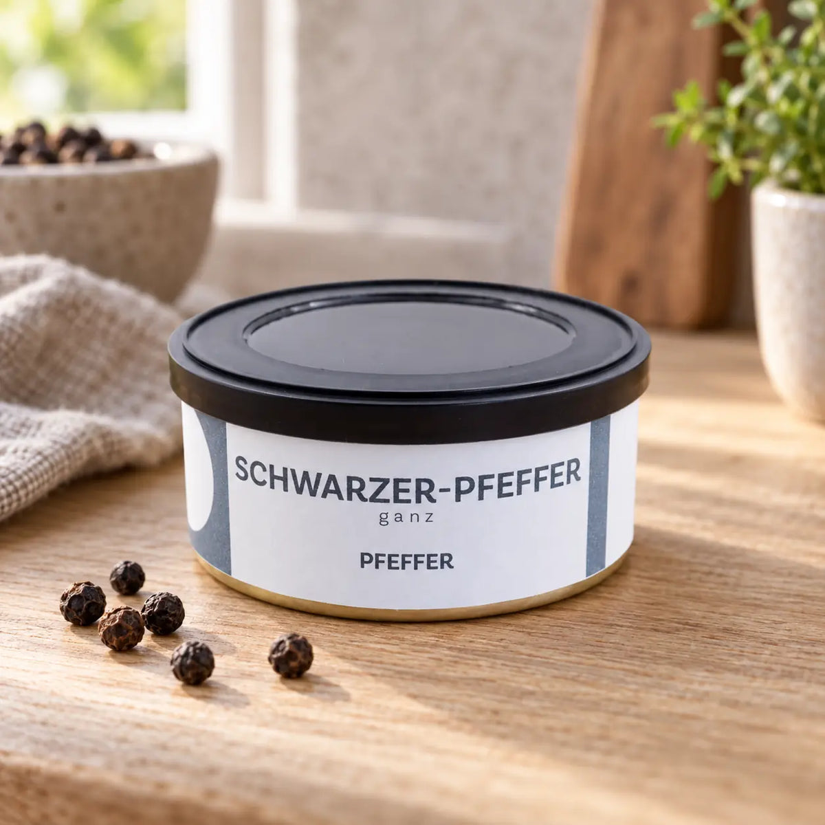 Schwarzer Pfeffer ganz in der originalen flachen Dose mit hellblau-weißem Etikett neben einzelnen Pfefferkörnern.