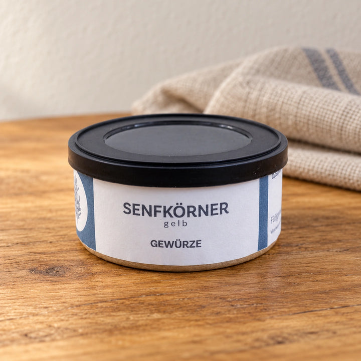 Originaldose Senfkörner gelb auf einer warmen Holzfläche neben einem Leinentuch.