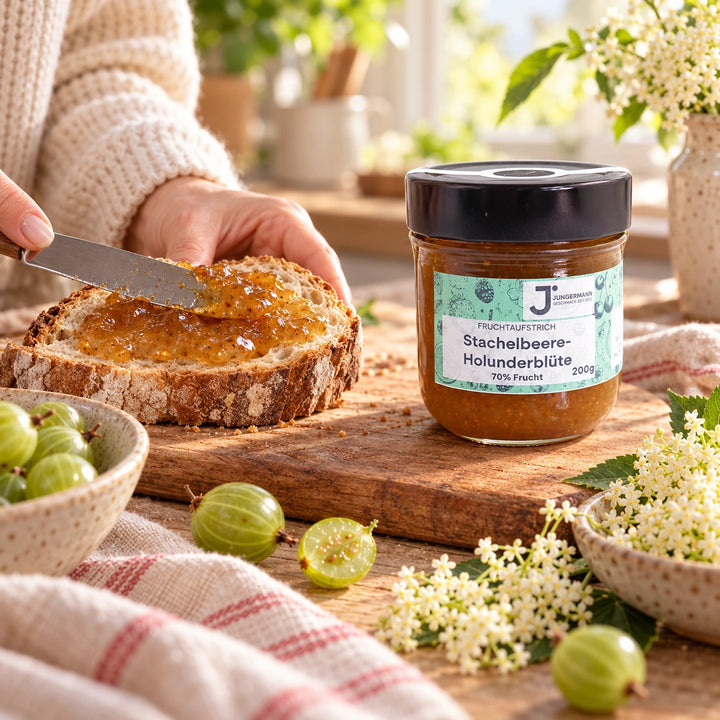 Eine Hand streicht Stachelbeer-Holunderblüten Fruchtaufstrich auf eine Scheibe Bauernbrot, neben dem Originalglas und grünen Stachelbeeren