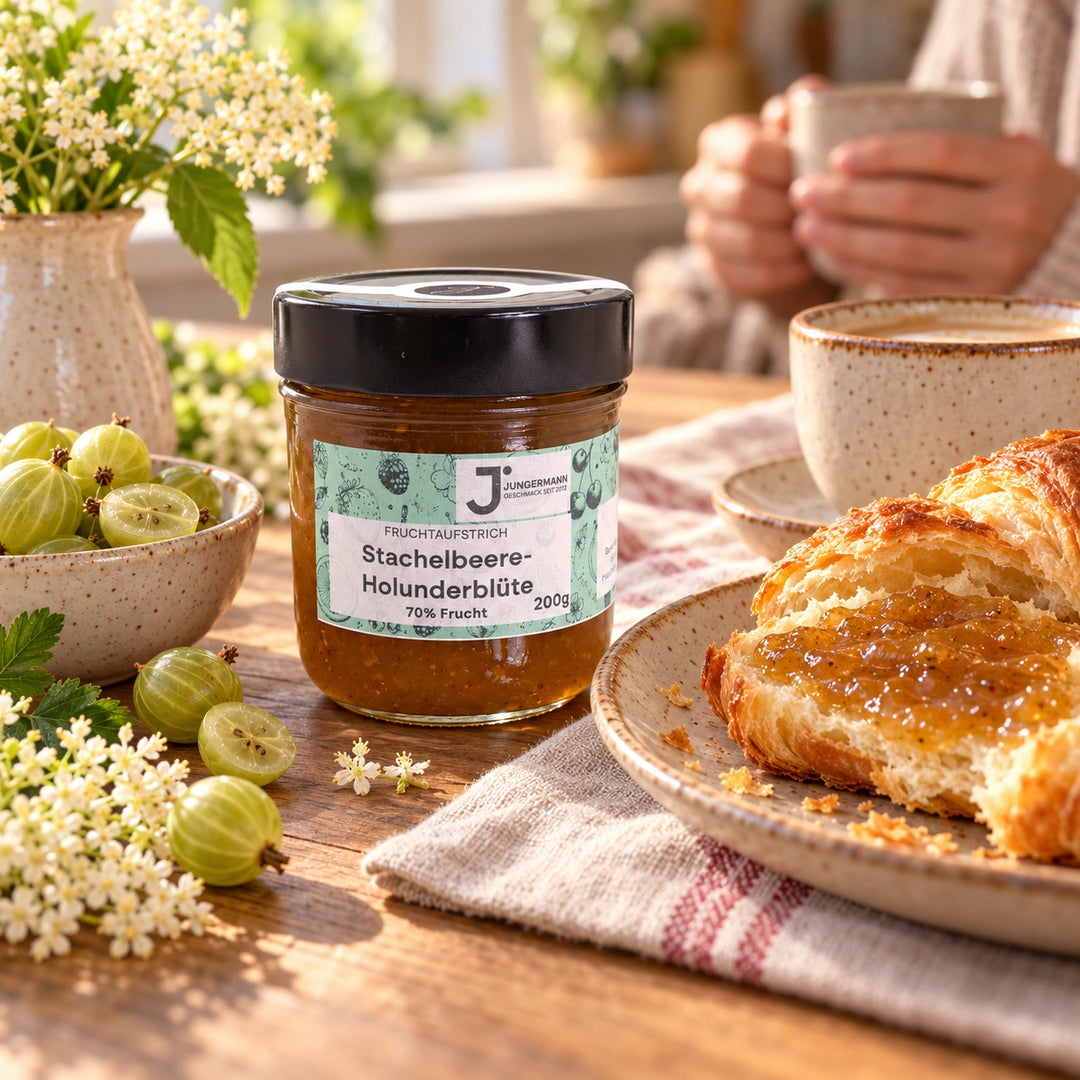 Frühstückstisch mit Stachelbeer-Holunderblüten Fruchtaufstrich, bestrichenem Buttercroissant, Kaffee und grünen Stachelbeeren